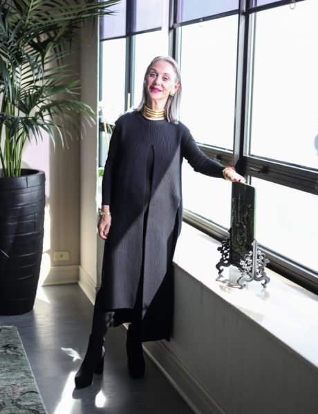 Honey Good showing how to wear a sweater in a black duster-length sweater standing by a window.