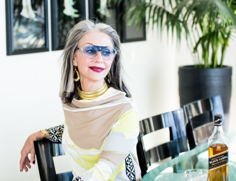 Image of a woman with gray hair looking over her shoulder seated at a table, with a rye smile and red lipstick knowing how to find strength in the unknown.