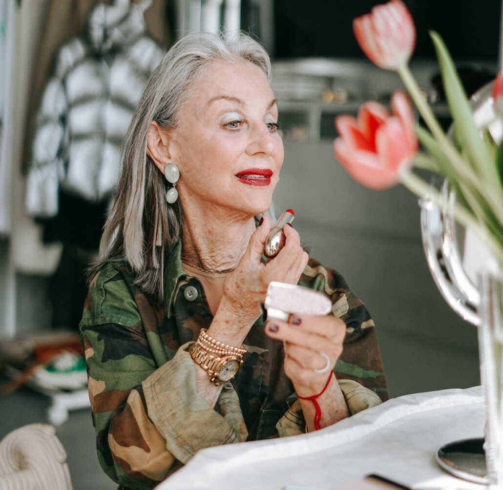 Honey Good puts on bold red lipstick after 50 sitting at her vanity holding one of her favorite red lipsticks for women over 50. She's wearing a camo shirt with rolled-up sleeves, a gold watch, and gold bracelets.