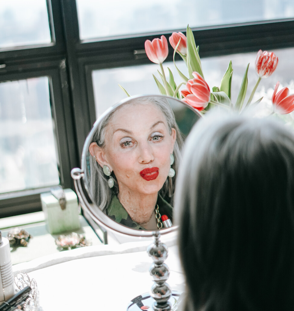 honeys-skincare-routine honey good wearing red lipstick at her vanity and her reflection is shown in a round mirror with a window in the background. She loves to explore the best red lipsticks for women over 50