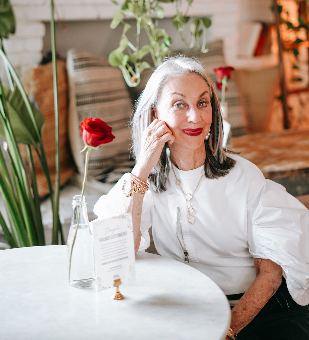 Image: Honey Good sitting at cafe table, head propped on hand with a single rose on table contemplating rose-colored glasses attitute