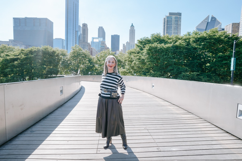 Image: Honey Good showing how to wear a belt bag posing on long walkway, wearing striped shirt and black leather skirt with Chicago skyline in the background.