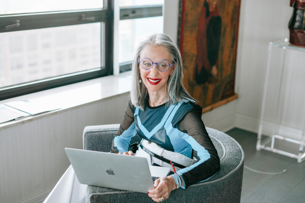 Honey Good at a chair by the window overlooking the city with her laptop in her lap, smiling at the screen showing why you should consider journaling.